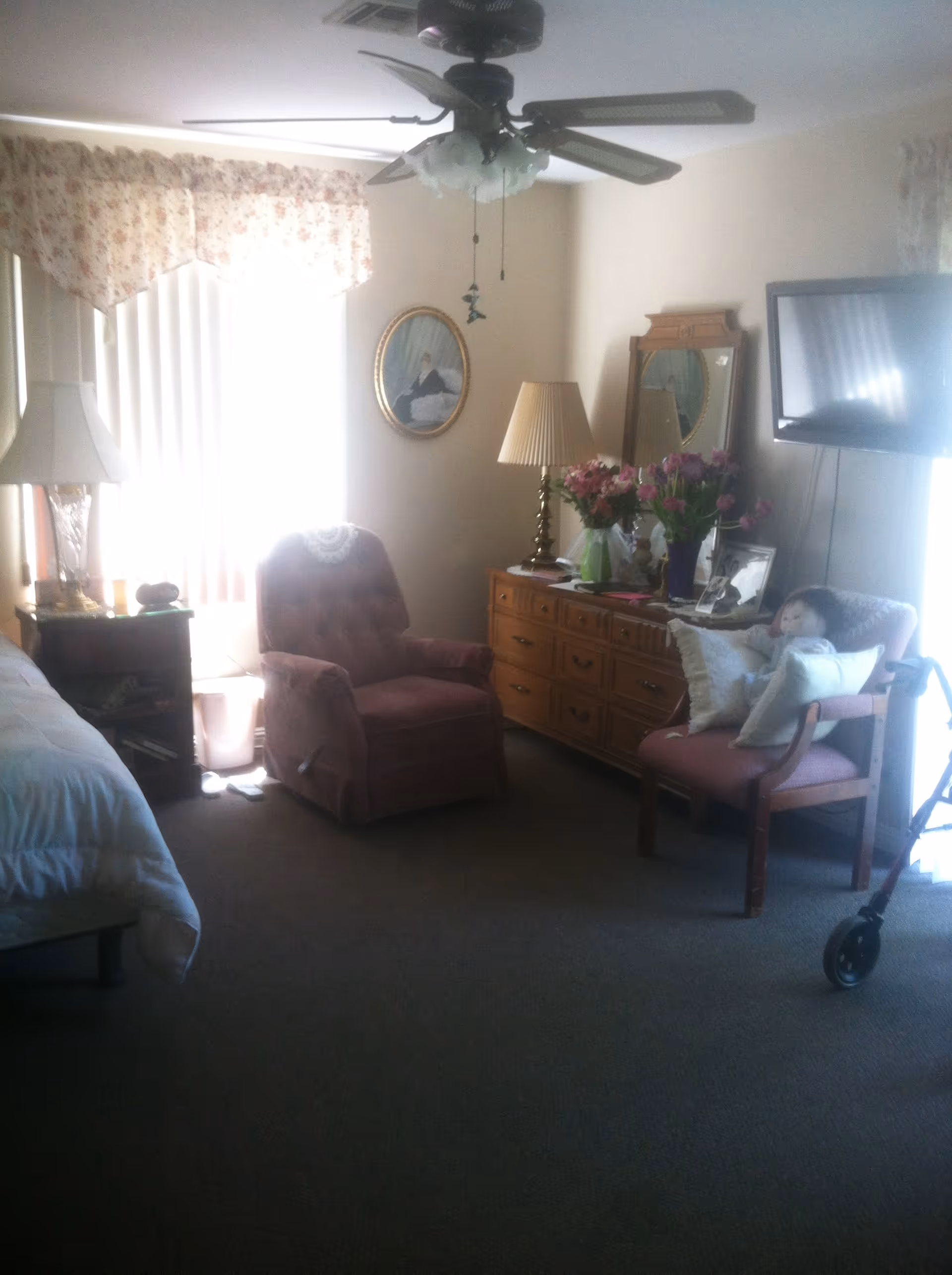 A cozy bedroom with a bed partially visible on the left, a pink armchair in front of a window with floral curtains, a wooden dresser with a lamp, flowers, and framed photos, a wall-mounted TV, and another chair with pillows and a doll. A ceiling fan is mounted above.