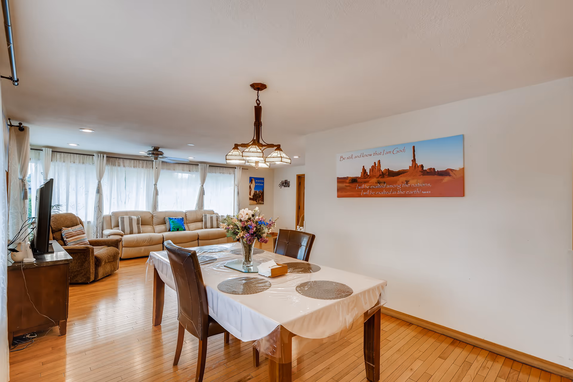 Open-plan living area with a dining table and chairs in the foreground and sofas, a TV, and large curtained windows in the background.