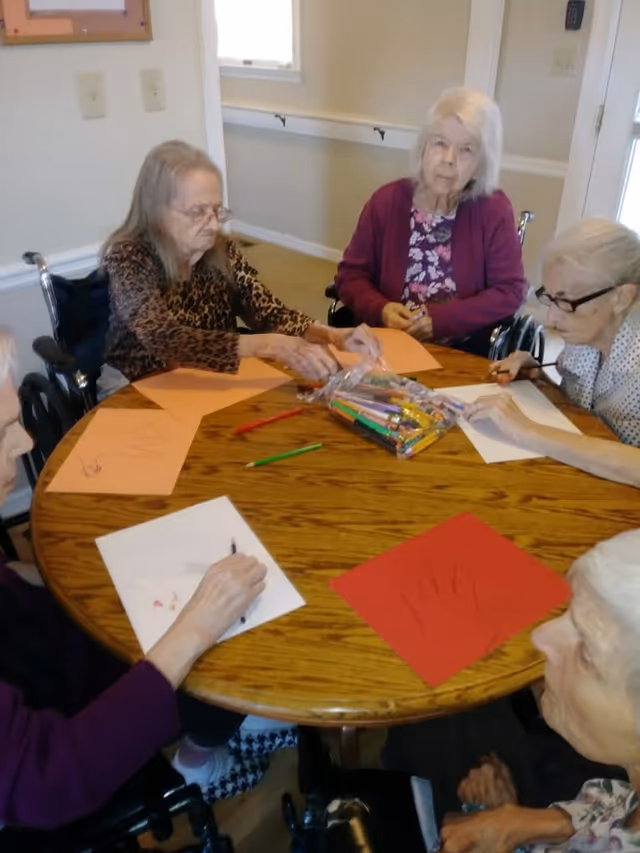 Five elderly women sitting around a wooden round table engaged in an arts and crafts activity with colored paper and markers. Some of the women are in wheelchairs, and the setting appears to be a well-lit room with light-colored walls and a window in the background.