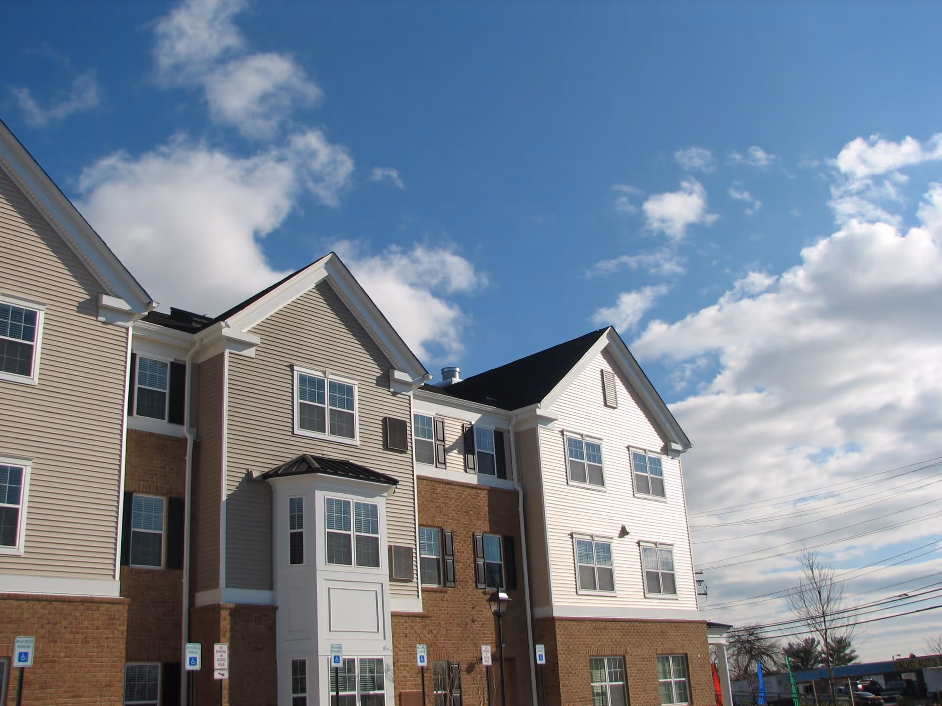 Exterior view of a multi-story senior apartment building with beige and white siding, brick accents, multiple windows, and a clear blue sky with some clouds in the background.