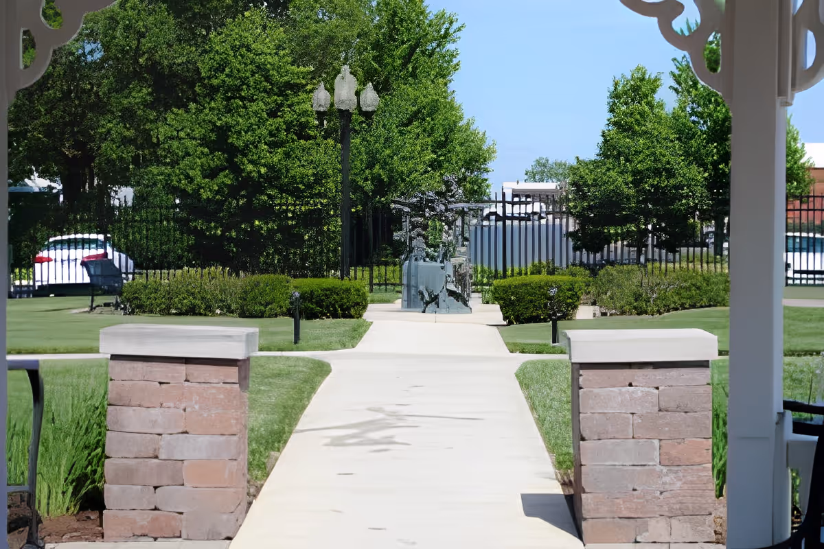 View of a paved walkway leading through a well-maintained garden area with green grass, bushes, and trees. The walkway is flanked by two stone pillars and leads to a black metal fence with a gate. There is a decorative white structure partially visible on the right side of the image and a streetlamp in the background.