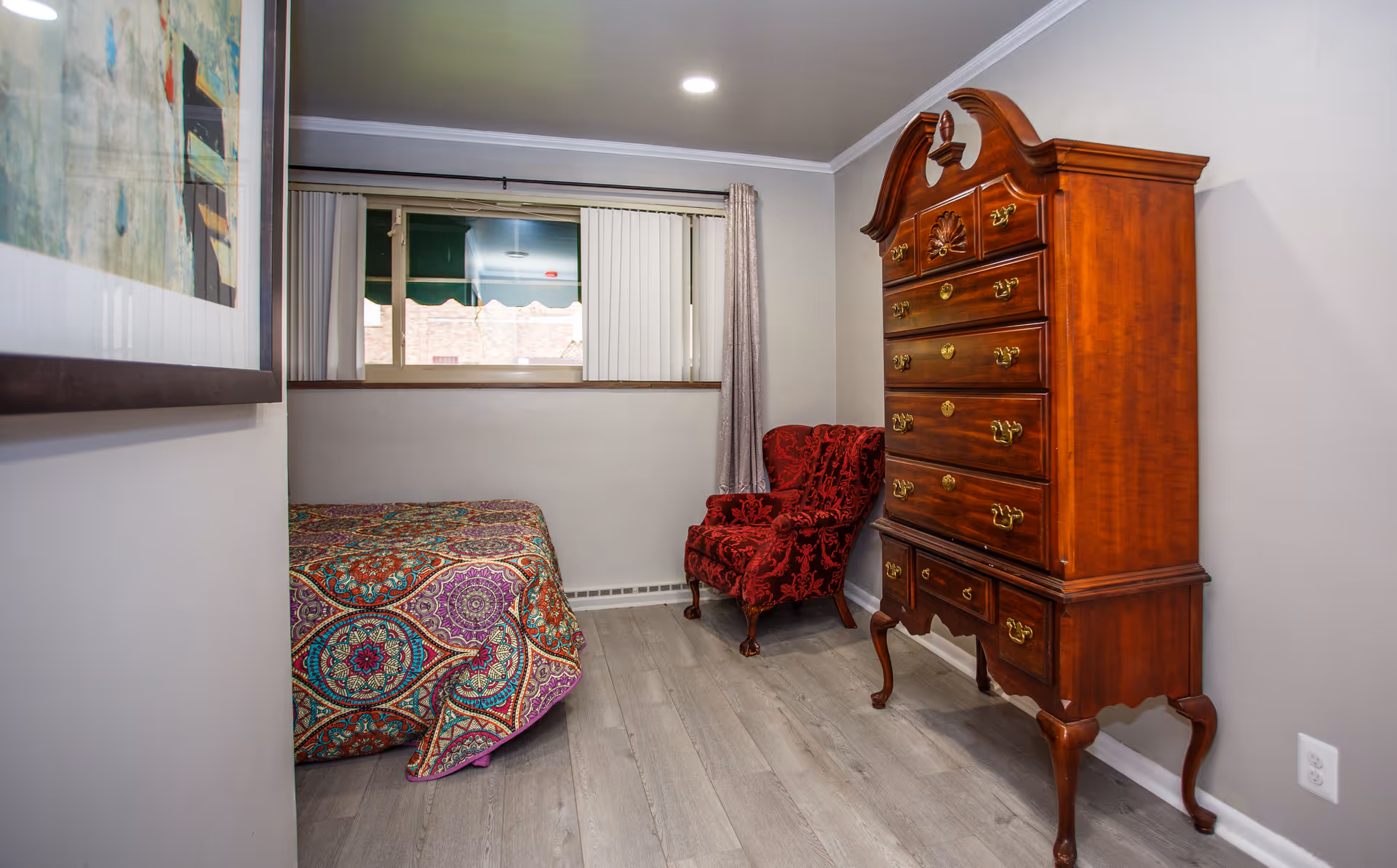 A small bedroom with a colorful patterned bedspread on the bed, a red upholstered armchair, and a tall wooden dresser with multiple drawers. There is a window with vertical blinds and a curtain, and a framed picture on the wall.