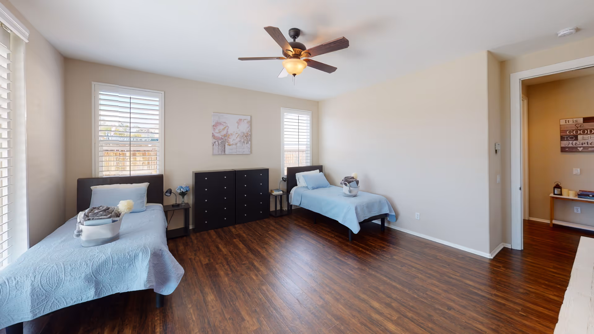 Spacious bedroom with two twin beds, a dark dresser between windows, hardwood floors and a ceiling fan.