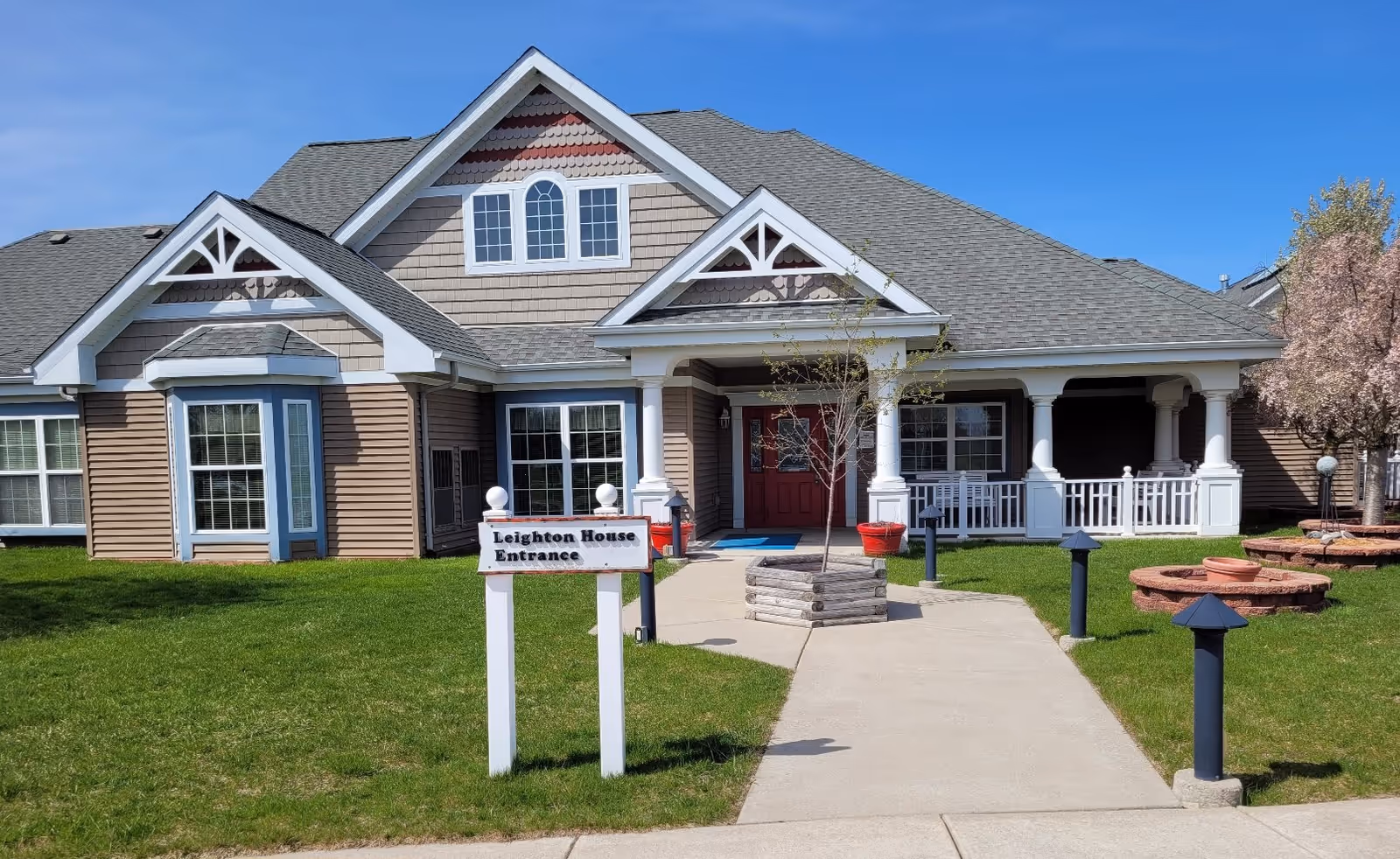Exterior view of a single-story building with beige and brown siding, white trim, and a gray shingled roof under a clear blue sky. A concrete walkway leads to a covered entrance with white columns and a red door. There is a sign in front that reads 'Leighton House Entrance' and a small tree planted in a wooden planter near the walkway. The lawn is green and well-maintained with some outdoor lighting fixtures and a circular brick planter on the right side.