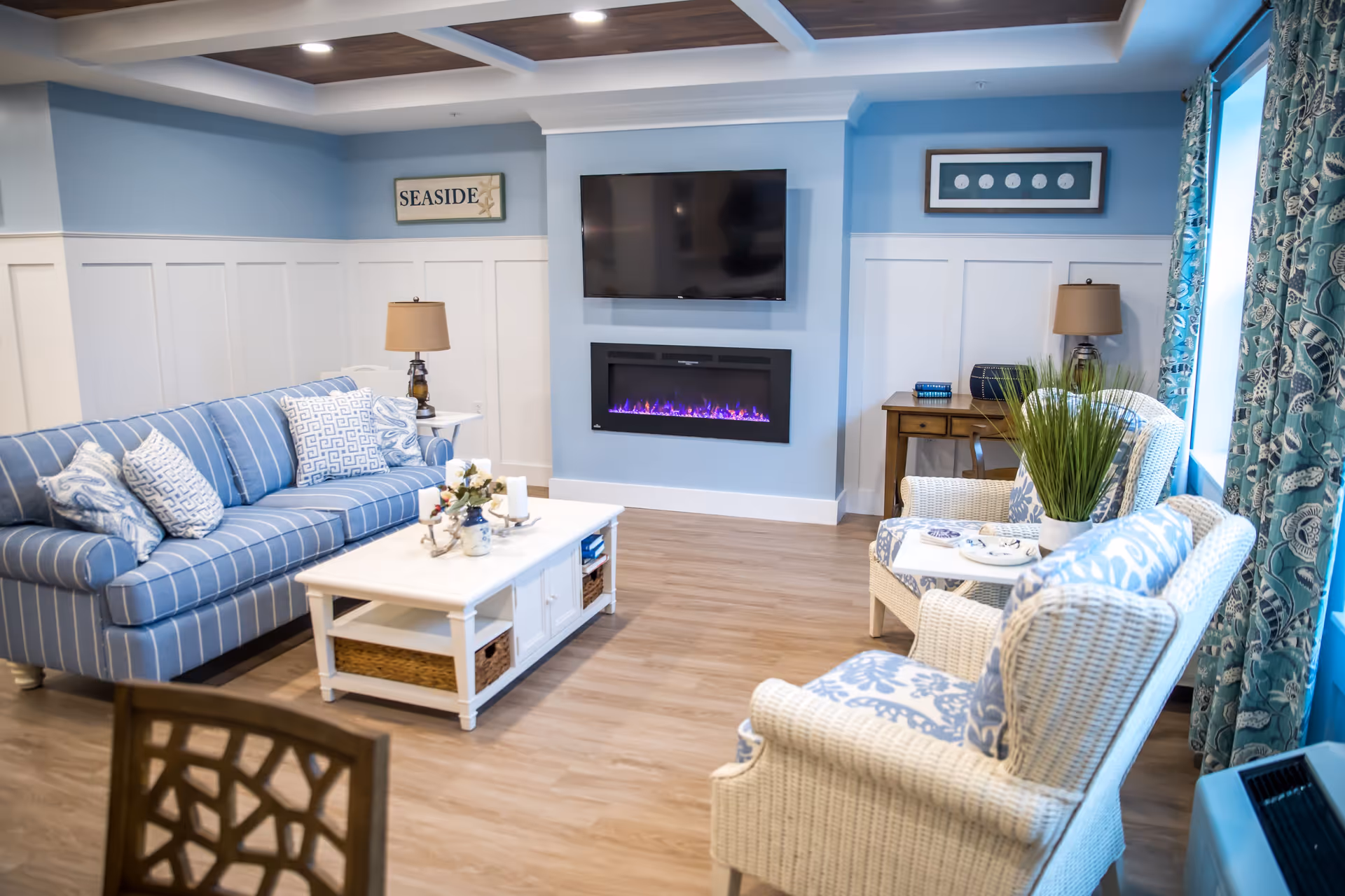 A cozy living room with a blue and white color scheme featuring a striped blue sofa with patterned pillows, two white wicker armchairs with blue patterned cushions, a white coffee table with decorative items, a wall-mounted flat screen TV above a modern electric fireplace with purple flames, two table lamps on side tables, and blue patterned curtains on the window.