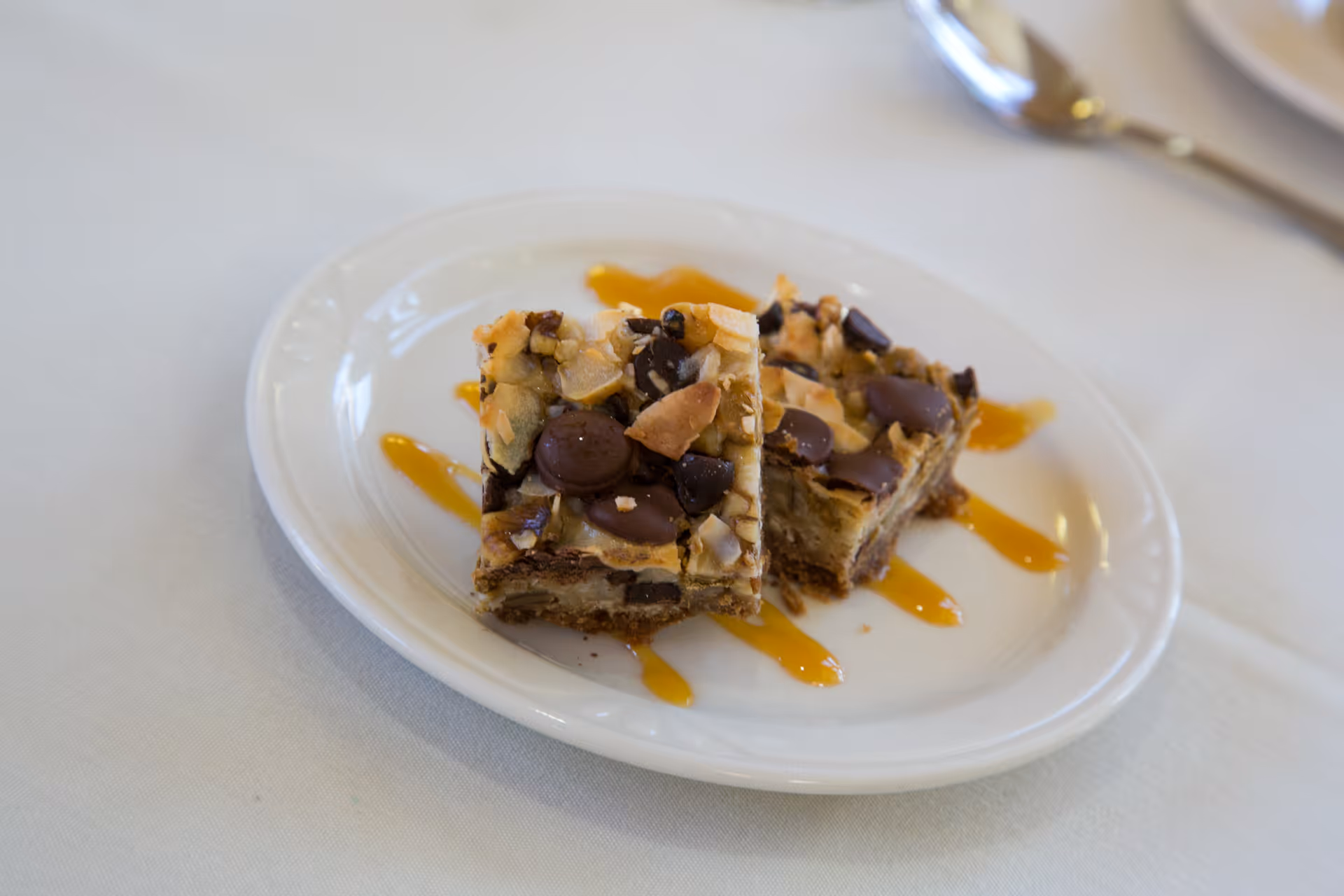 Two dessert bars with chocolate chips and nuts on a white plate drizzled with caramel sauce, placed on a white tablecloth with a spoon in the background.