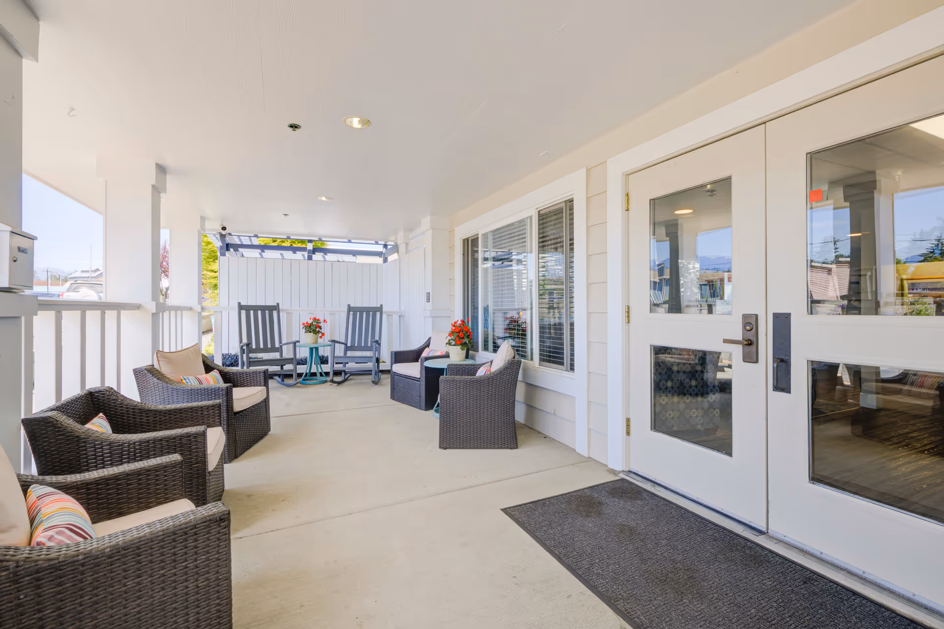 Covered outdoor patio area with wicker chairs and cushions, two rocking chairs, small tables with potted flowers, and double glass doors leading inside.