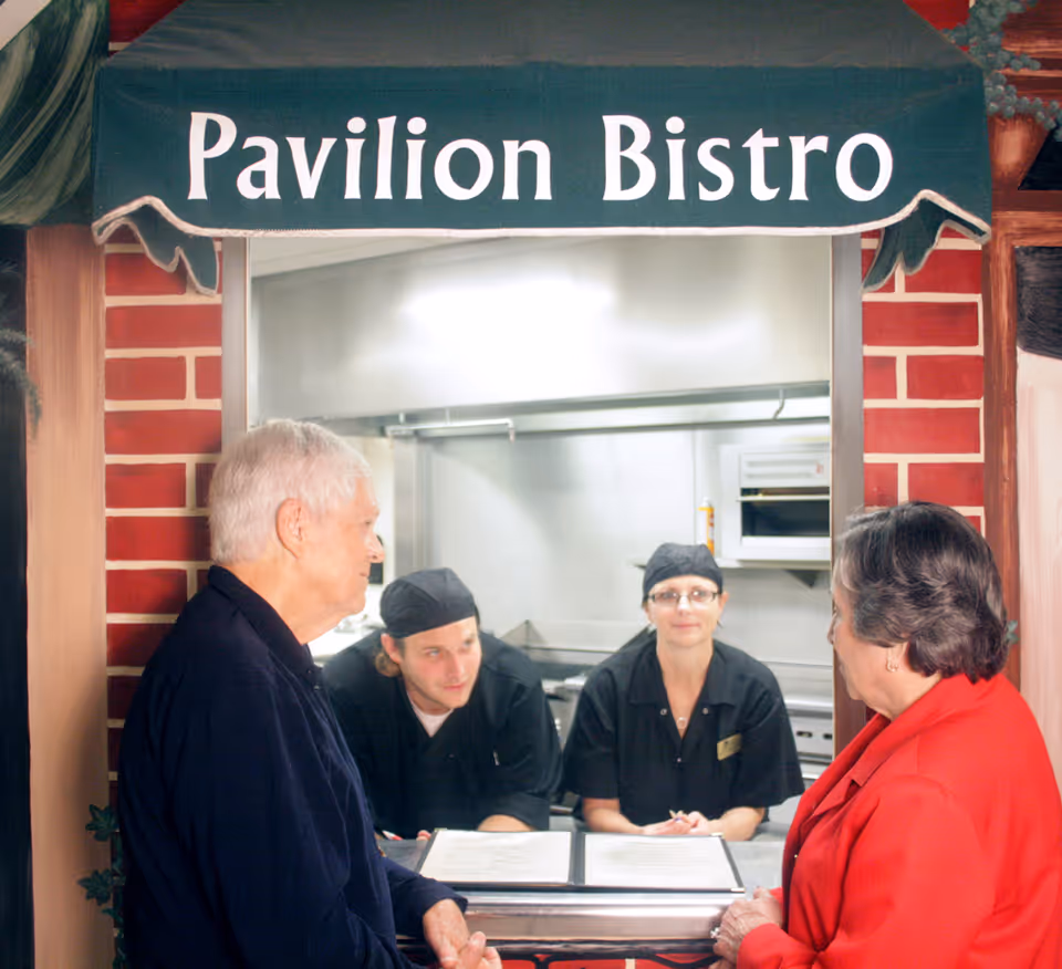 Two elderly residents speak with kitchen staff at the Pavilion Bistro service window under a green awning.