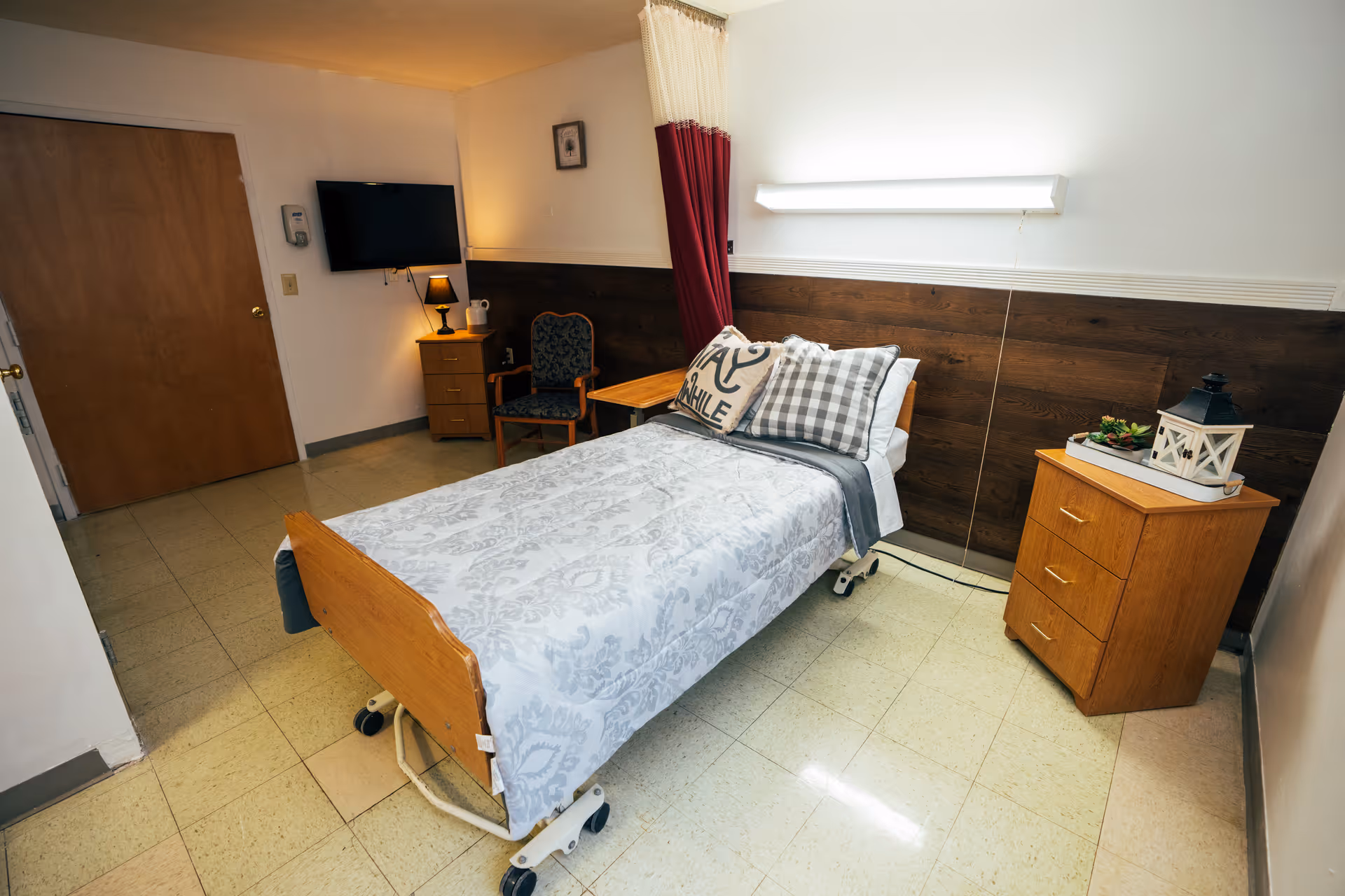 A single hospital-style bed with patterned bedding and pillows in a small, clean room. The room has a wooden headboard, a bedside table with a lantern and plant, a chair, a small wooden dresser with a lamp, and a wall-mounted TV. The floor is tiled, and there is a curtain that can be drawn around the bed for privacy.