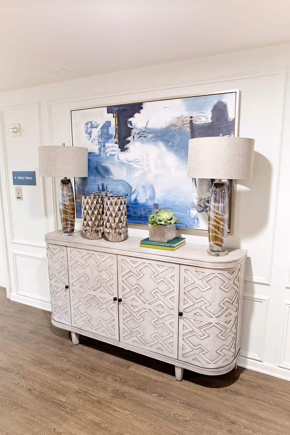 A decorative console table with a geometric pattern on the doors, topped with two tall table lamps with beige shades, two textured vases, and a small potted succulent plant. Above the table hangs a large abstract painting in shades of blue, white, and black. The setting appears to be a hallway with wood flooring and white paneled walls, with a sign indicating the West Wing.