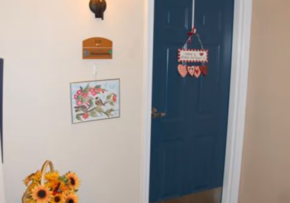 A hallway corner in a senior living facility with a dark blue door decorated with a hanging sign featuring hearts. Next to the door is a wall with a small wooden plaque and a framed picture of birds on a branch with pink flowers. Below the picture is a basket filled with bright yellow sunflowers.