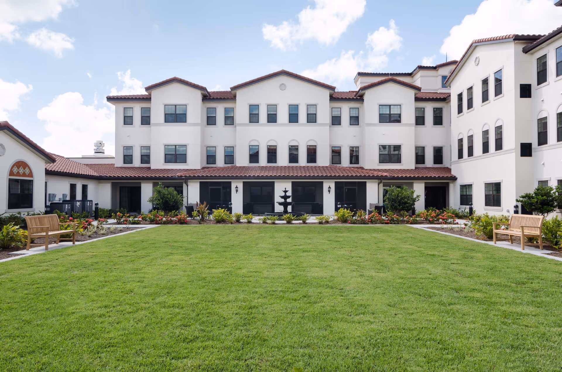 A well-maintained green lawn with flower beds and shrubs in front of a large, white multi-story building with a red-tiled roof under a partly cloudy sky. There are two wooden benches on either side of the lawn and a black fountain in the center near the building.