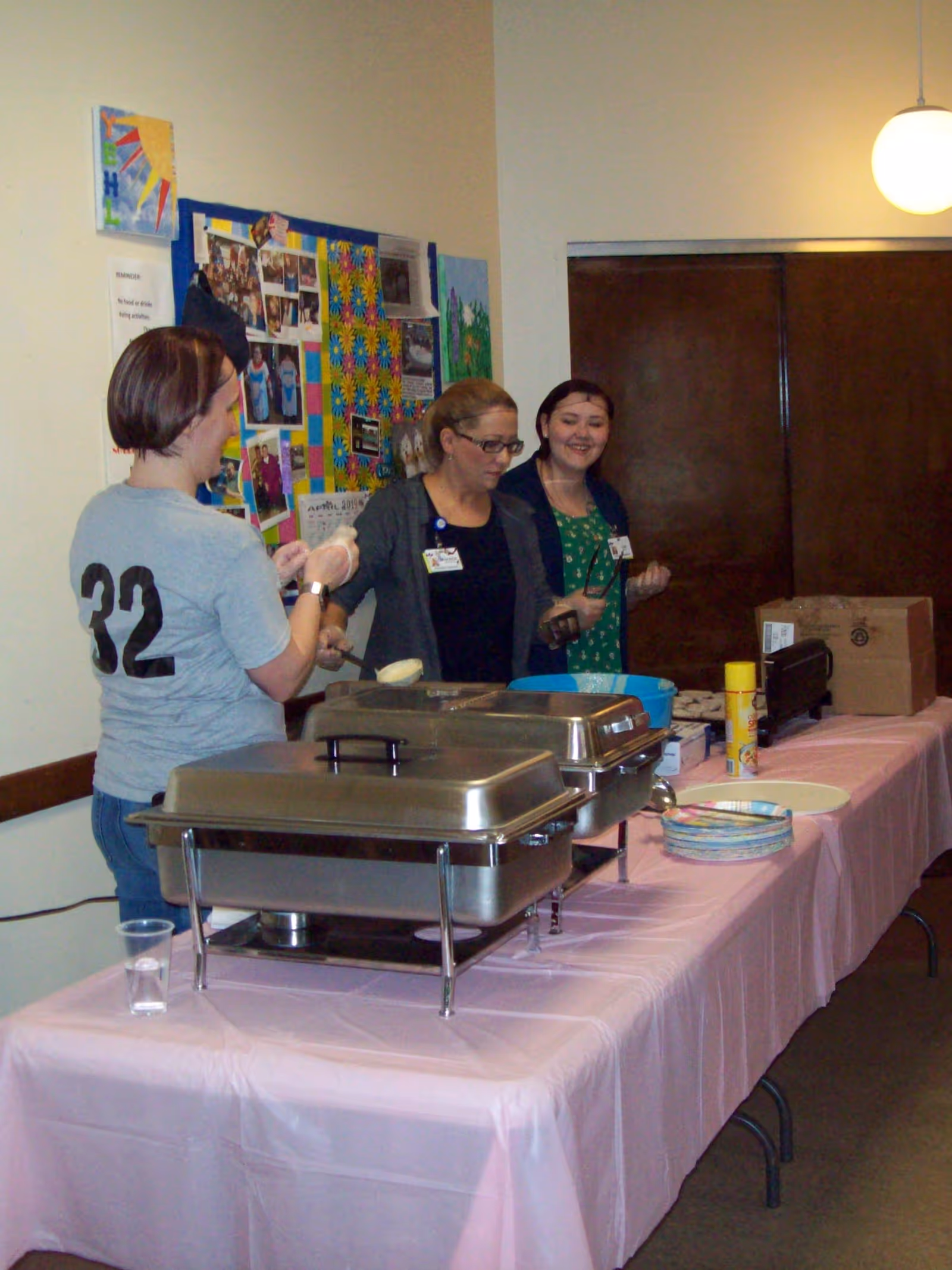 Three women standing behind a table covered with a pink tablecloth, serving food from chafing dishes in a room with a bulletin board displaying colorful photos and artwork on the wall.