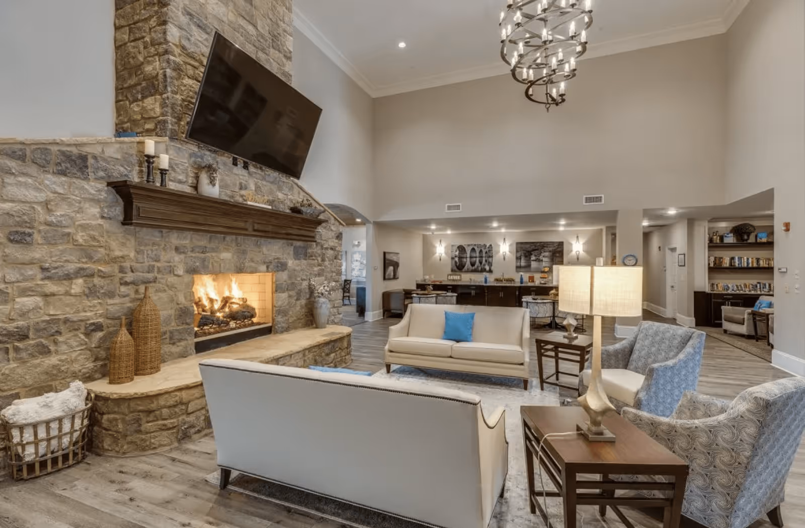 A spacious and well-lit living room area in an assisted living facility featuring a large stone fireplace with a mounted flat-screen TV above it. The room is furnished with cream-colored sofas, patterned armchairs, a wooden side table with a lamp, and decorative items including woven baskets and candles. The background shows a kitchenette or refreshment area with bar stools and wall art.