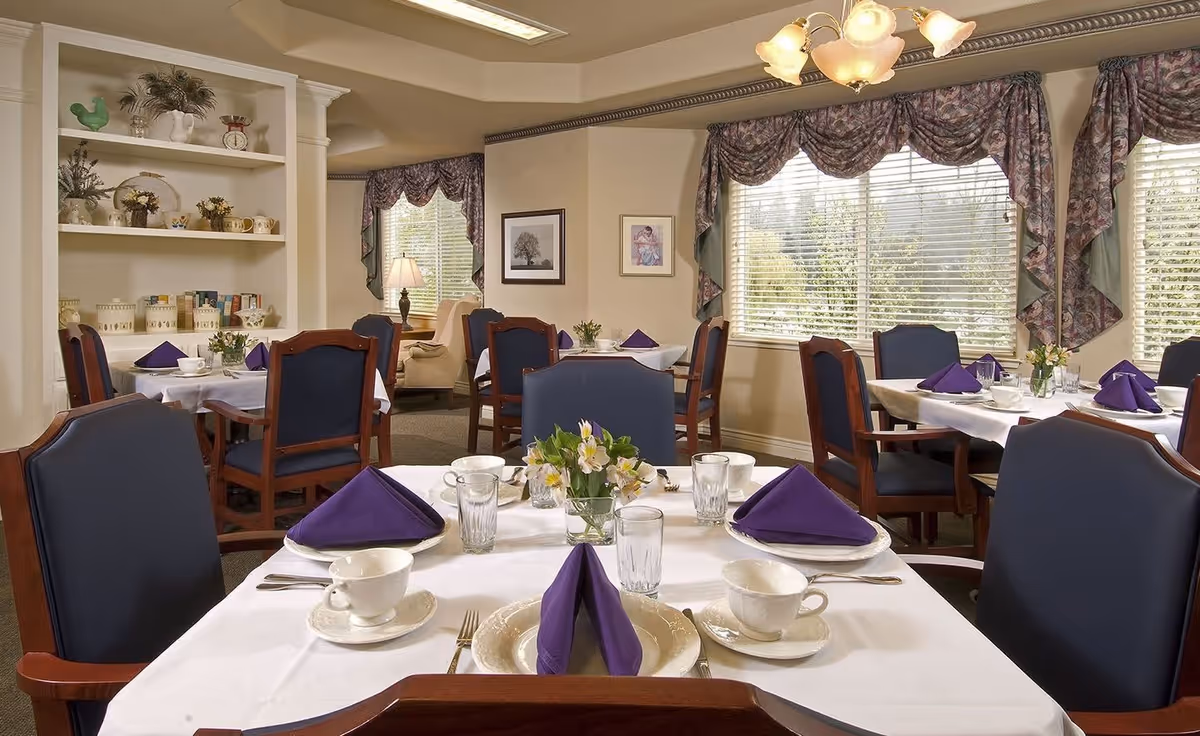 A dining room with several tables set with white tablecloths, white plates, cups, glasses, silverware, and purple folded napkins. The room has large windows with floral curtains, wooden chairs with dark blue cushions, and a chandelier overhead. There is a built-in shelf with decorative items and framed pictures on the walls.