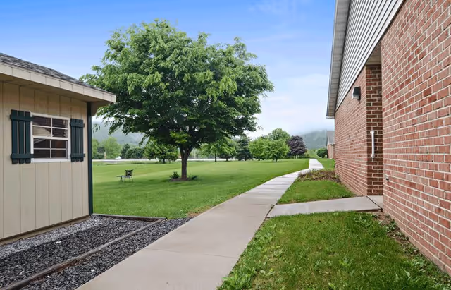 A paved walkway runs between a brick building on the right and a small beige shed with green shutters on the left. The walkway leads into a grassy area with several trees and a bench under one of the trees. The sky is clear with some clouds.