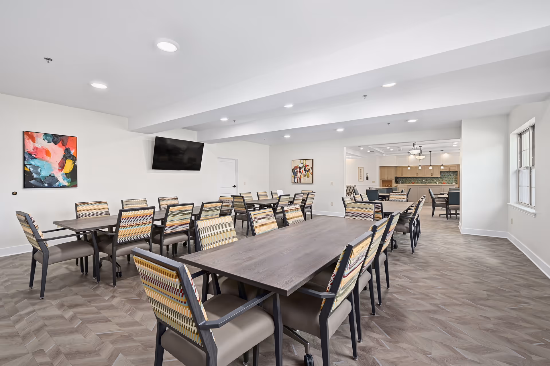 A spacious dining room with multiple rectangular tables and patterned chairs arranged neatly. The room has light-colored walls, a mounted flat-screen TV, colorful abstract paintings, and large windows allowing natural light to enter. The flooring has a wood-like pattern, and the room extends into another area with additional seating and kitchen cabinetry visible in the background.