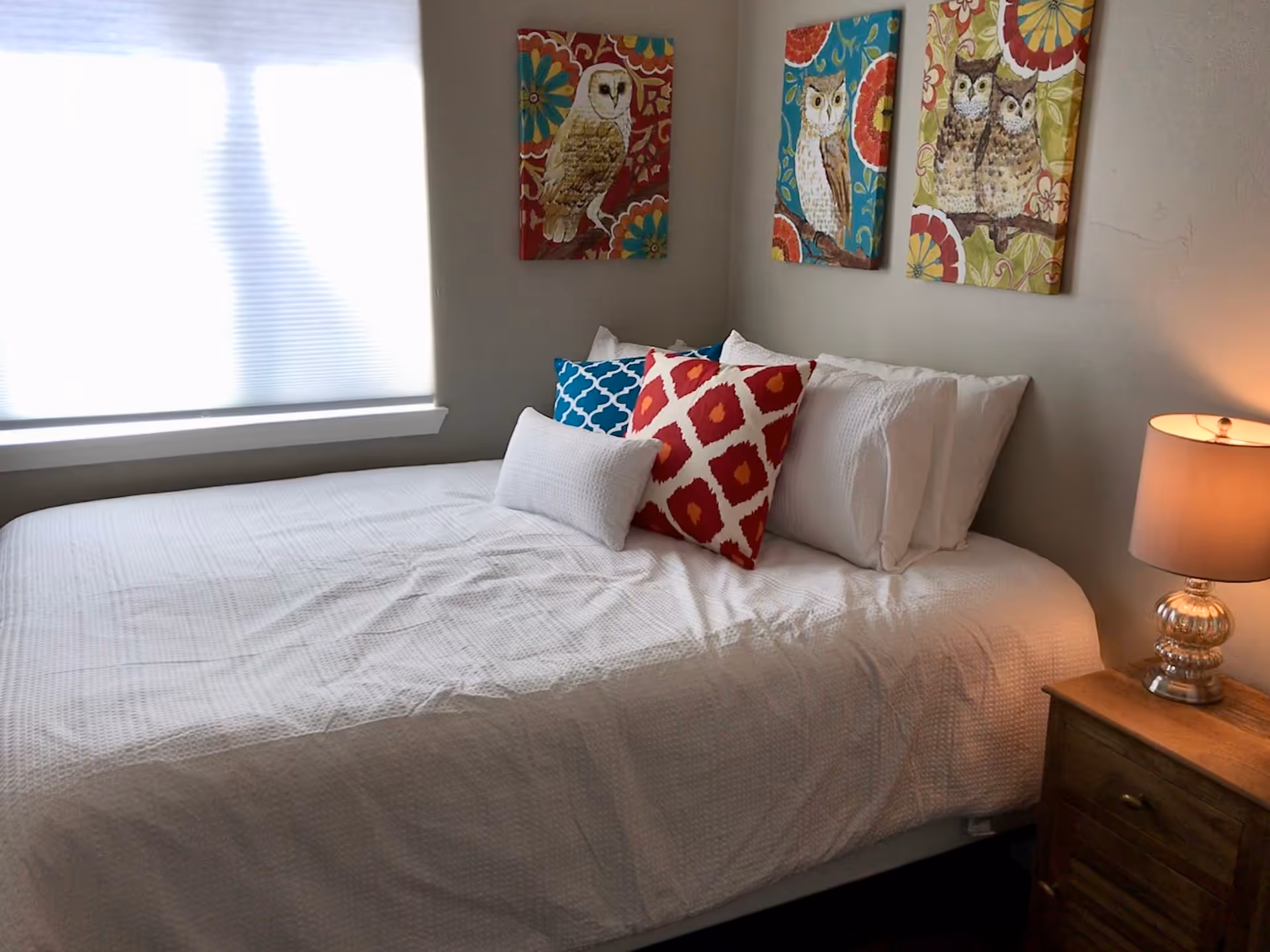 A neatly made bed with white bedding and colorful decorative pillows in red and blue patterns. Above the bed, there are three framed paintings of owls on a wall. To the right of the bed, there is a wooden nightstand with a decorative lamp. A window with a closed blind is on the left side of the bed.