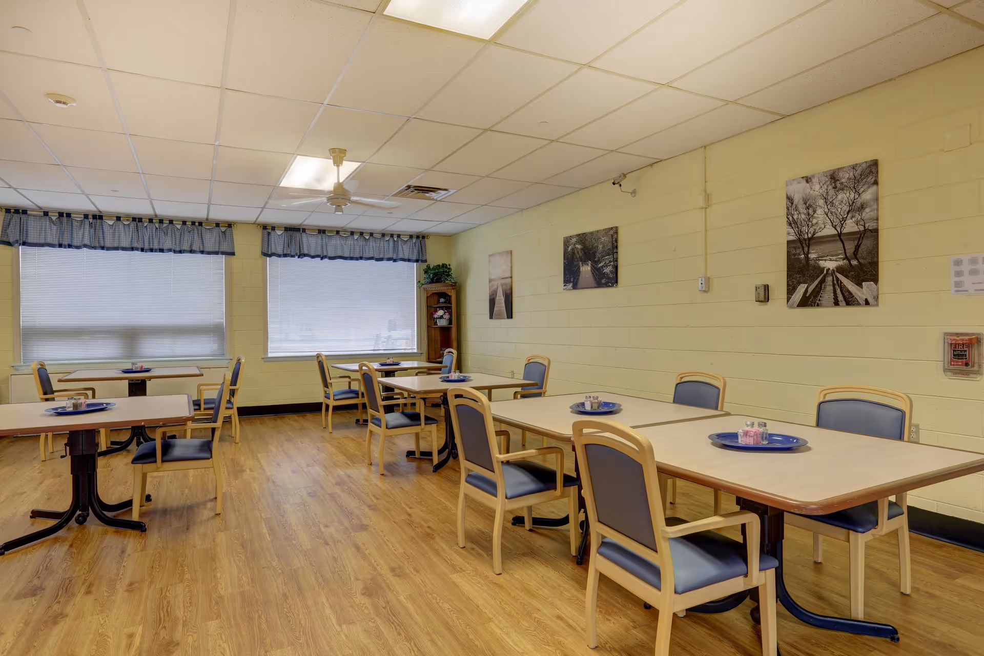 A dining room with several tables and chairs arranged neatly. Each table has a blue tray with condiments on it. The room has light yellow walls with framed pictures, large windows with blinds and valances, and a ceiling fan. The floor is wood-patterned.