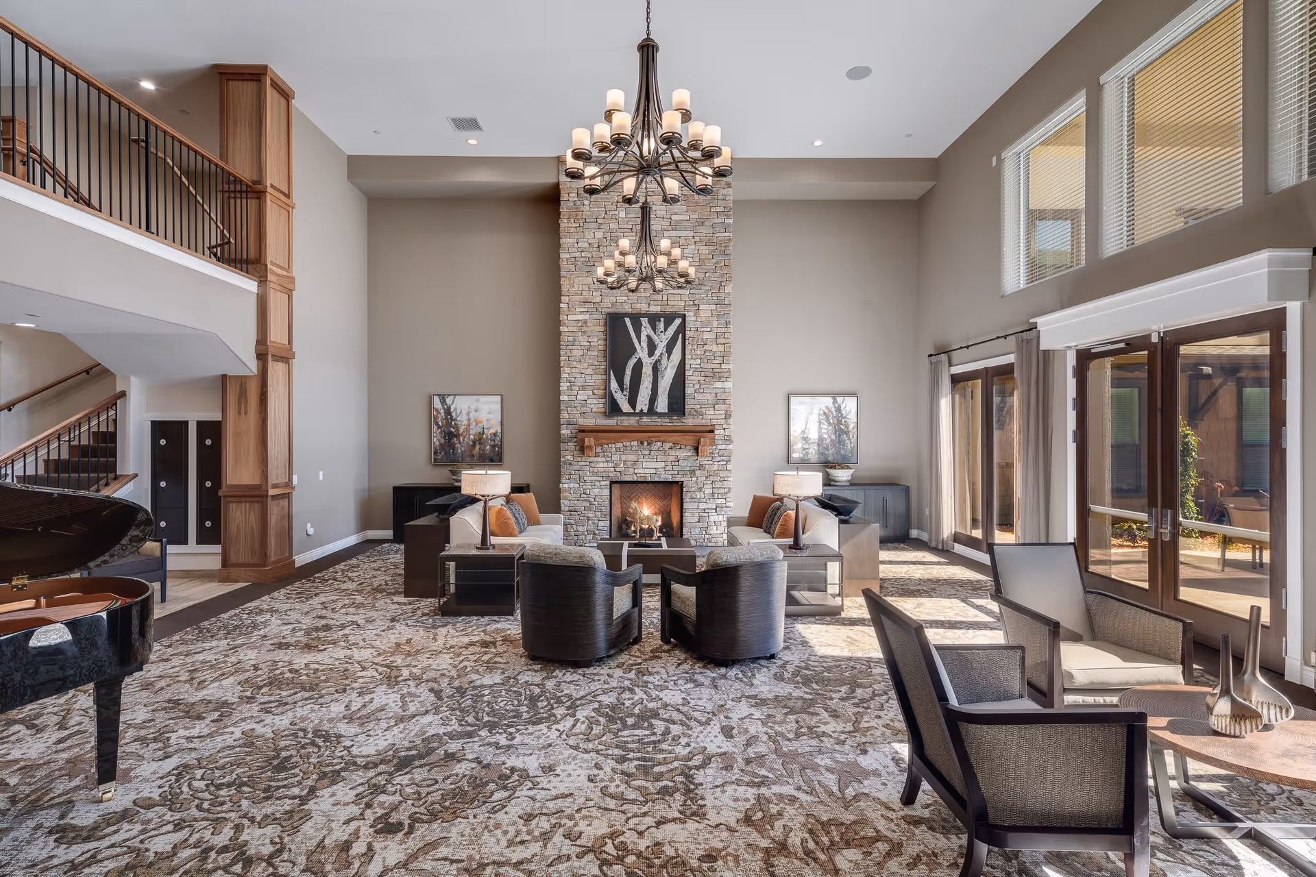 A spacious and elegant living room with high ceilings, a large stone fireplace in the center, two chandeliers, and large windows letting in natural light. The room is furnished with sofas, armchairs, side tables with lamps, a grand piano on the left, and a patterned carpet covering the floor.