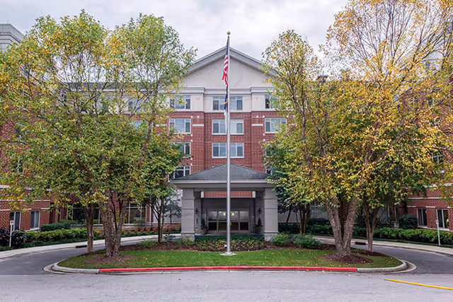Front exterior view of a multi-story brick building with a covered entrance, an American flag on a flagpole in the center, and trees with green and yellow leaves surrounding the entrance area.