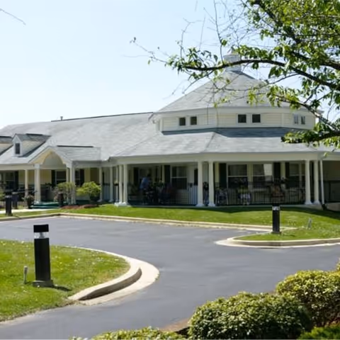 Exterior view of a single-story senior living facility building with a circular driveway, green lawn, and some bushes in the foreground. The building has a covered porch area with white columns and a gray roof.