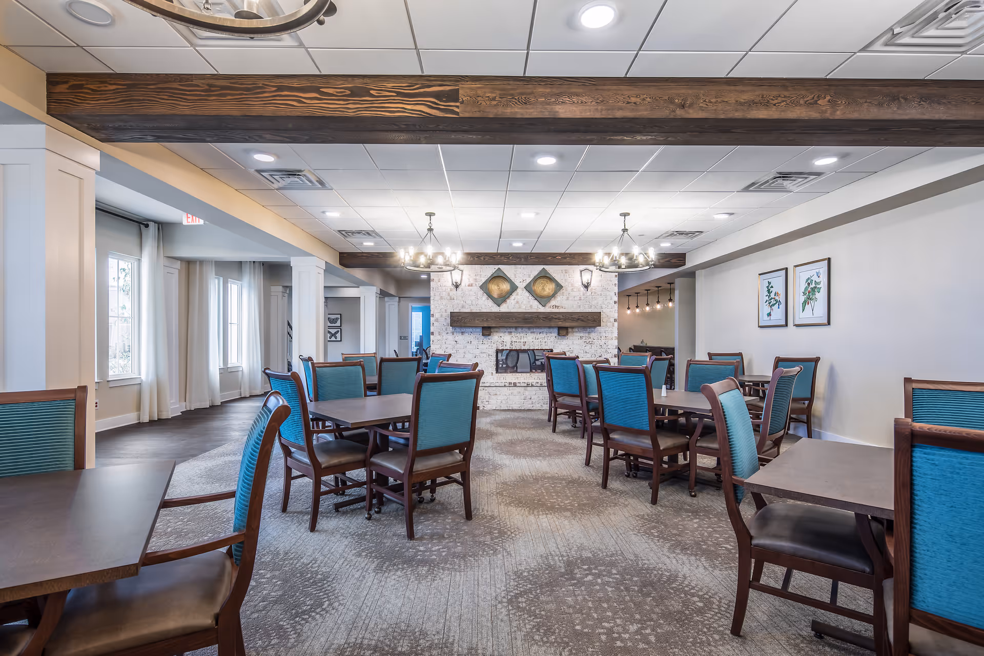 A spacious dining room with multiple wooden tables and chairs featuring teal upholstery. The room has a carpeted floor with a patterned design, a white brick fireplace with a wooden mantel, and two decorative wall hangings above the fireplace. The ceiling has recessed lighting and two chandeliers with candle-style bulbs. There are large windows with white curtains on the left side, and framed botanical prints on the right wall.