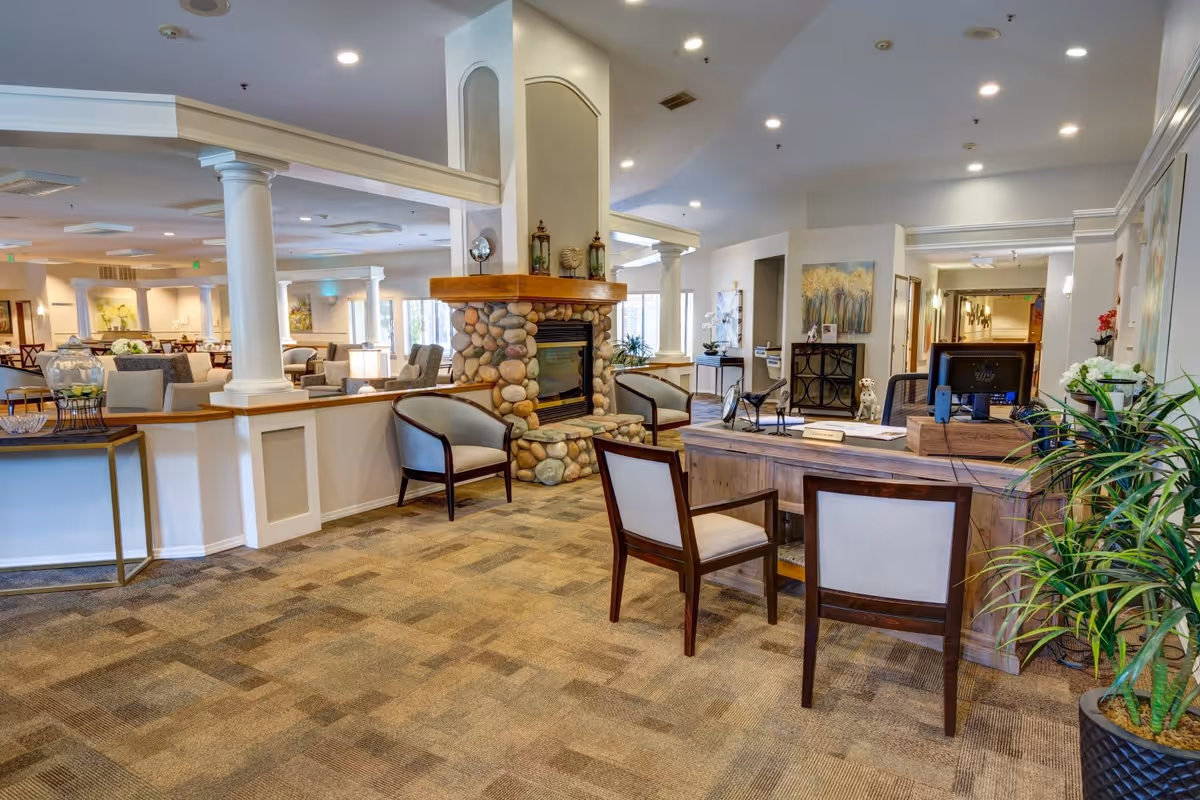 A spacious and well-lit senior living facility common area with a stone fireplace in the center, surrounded by comfortable chairs. There is a wooden reception desk with two chairs in front and a computer on top. The room features carpeted floors, white columns, and various plants and artwork on the walls.