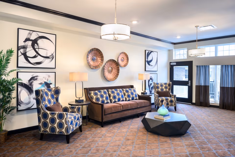 Well-lit communal living room with patterned armchairs, a sofa, wall art, lamps, and a geometric coffee table.