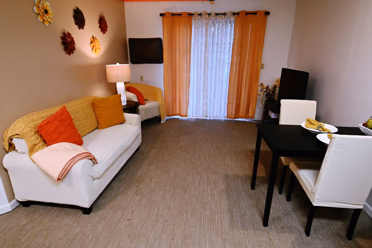 A cozy living room area with a white sofa adorned with orange and yellow cushions and a yellow throw blanket. There is a small side table with a lamp next to the sofa and an armchair with an orange cushion. Orange curtains and sheer white drapes cover a window or door at the back. A black dining table with two white chairs is set with plates and napkins on the right side of the room. The walls are decorated with flower-shaped wall hangings.