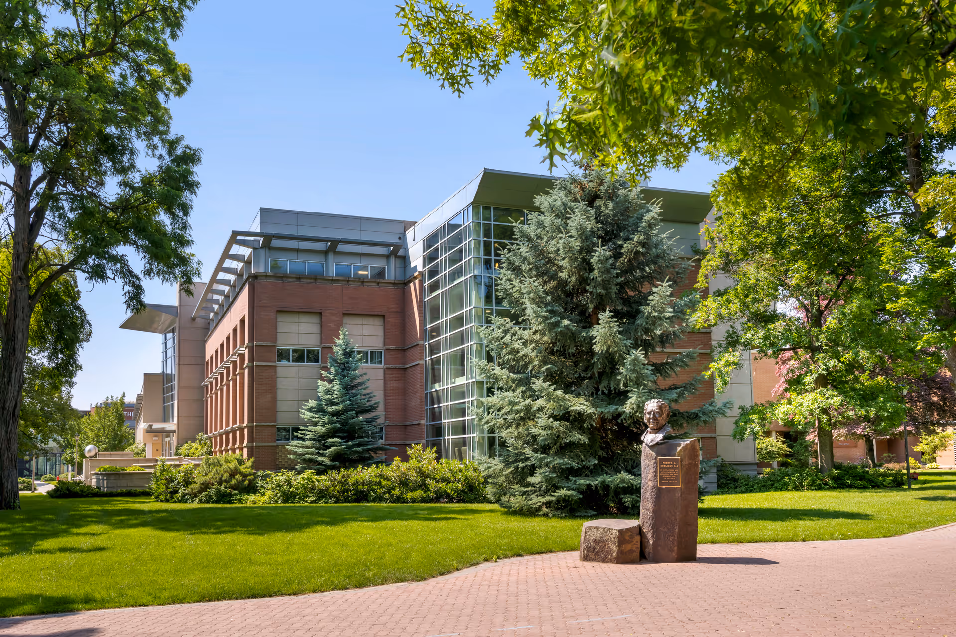 Exterior view of a modern brick and glass building surrounded by green trees and a well-maintained lawn. In front of the building, there is a statue bust mounted on a stone pedestal with a plaque. The sky is clear and blue.