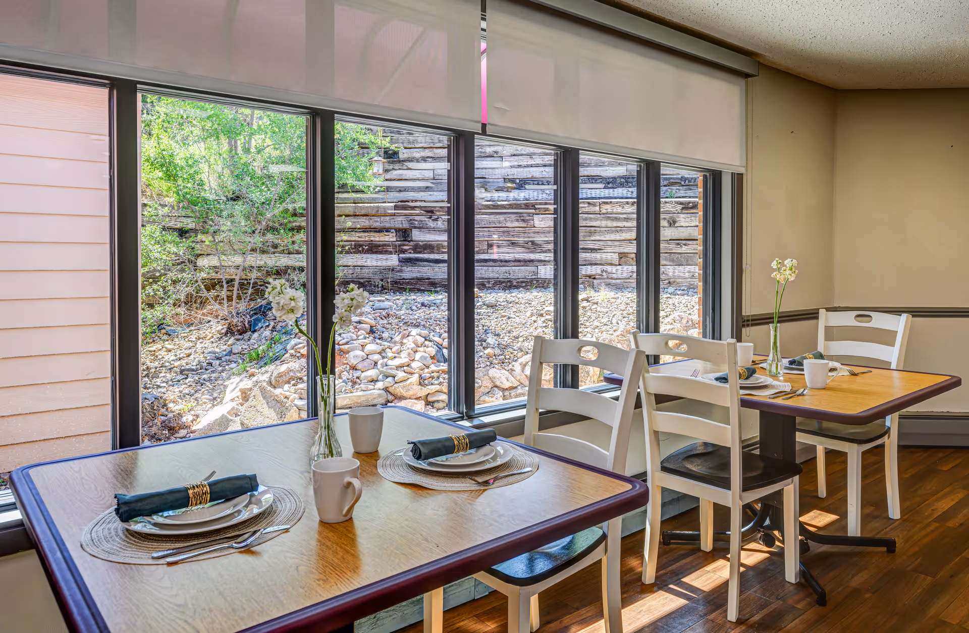 A dining area with two wooden tables set for two people each, featuring plates, napkins, mugs, and small vases with white flowers. Large windows behind the tables provide a view of a rocky outdoor area with greenery and a wooden retaining wall.