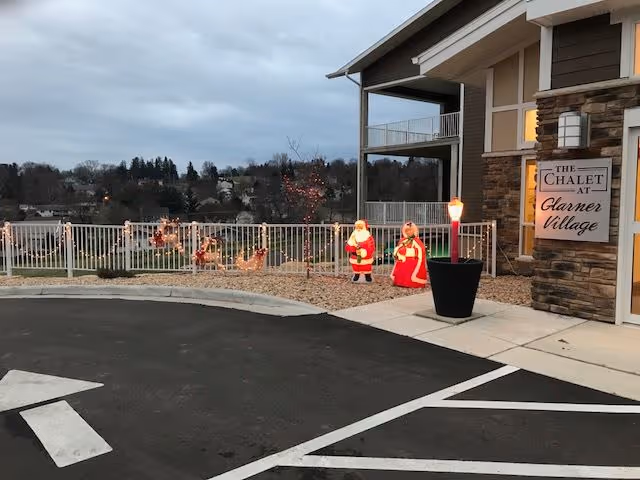 Outdoor view of a senior living facility entrance with holiday decorations including illuminated Santa Claus, Mrs. Claus, and reindeer figures near a white fence. The building has a sign that reads 'The Chalet at Glarner Village'. The sky is cloudy and it appears to be early evening.
