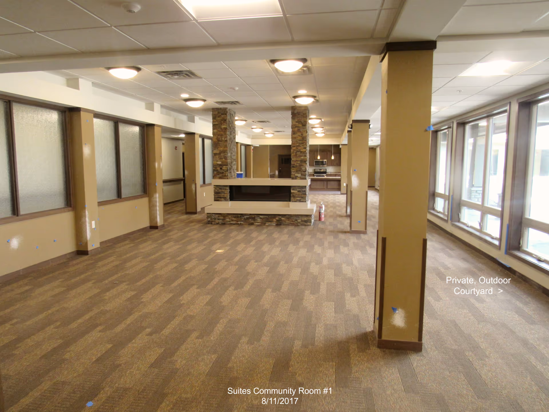 Spacious senior living community room with carpeted floor, support columns, large windows and a central stone fireplace/kitchen area.