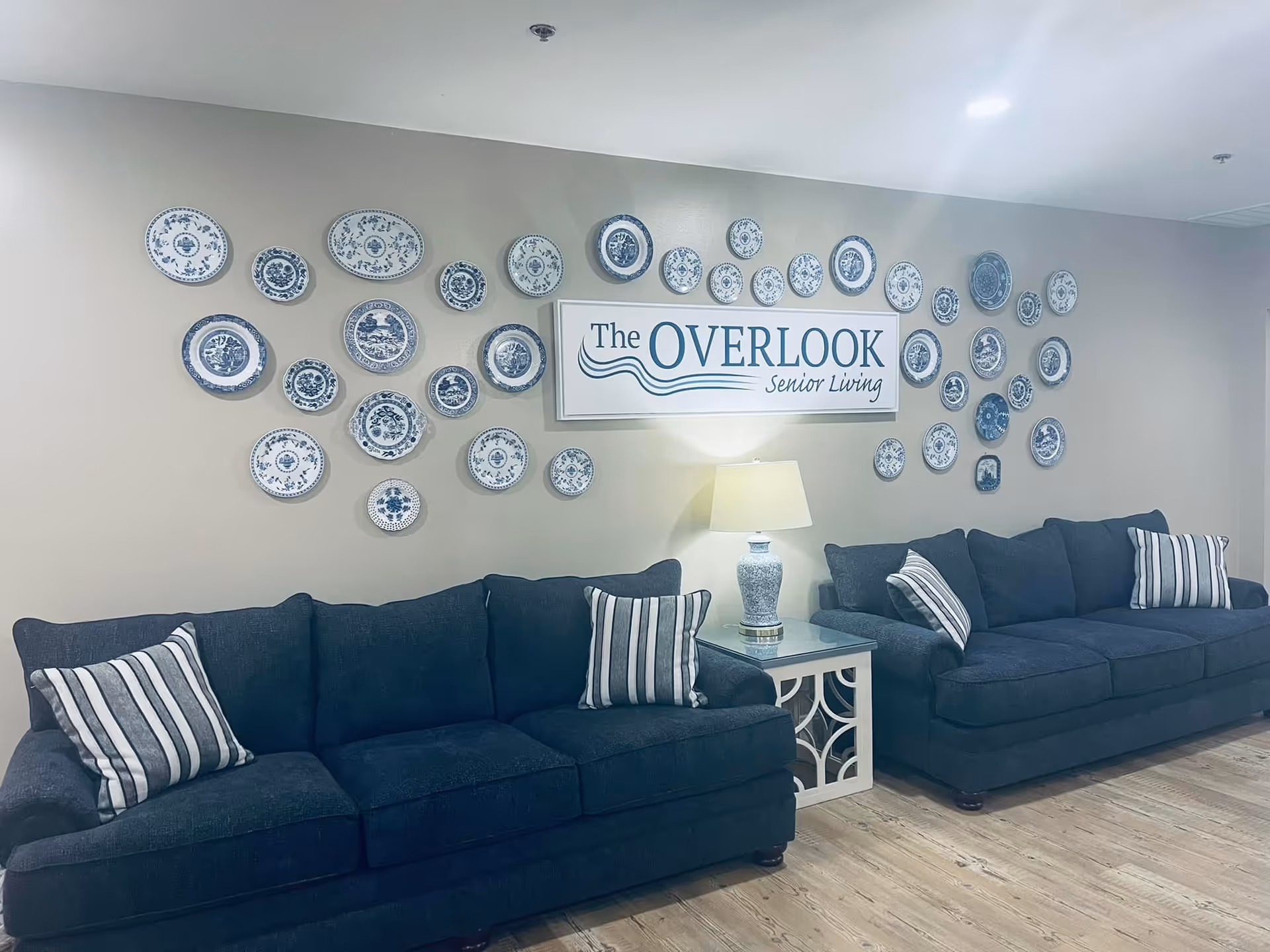 A cozy seating area with two dark blue sofas, each adorned with striped pillows. Between the sofas is a white side table with a decorative lamp. Above the table is a wall decorated with numerous blue and white decorative plates arranged around a sign that reads 'The Overlook Senior Living'. The floor is wooden, and the walls are painted a light beige color.
