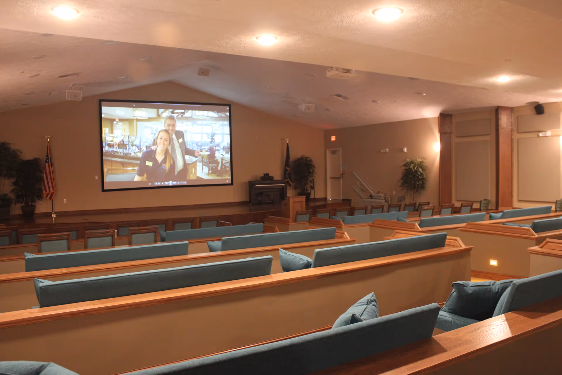 Interior view of a large room with multiple rows of wooden pew-style seating with blue cushions facing a large projection screen displaying two people. The room has beige walls, ceiling lights, an American flag, and some potted plants.