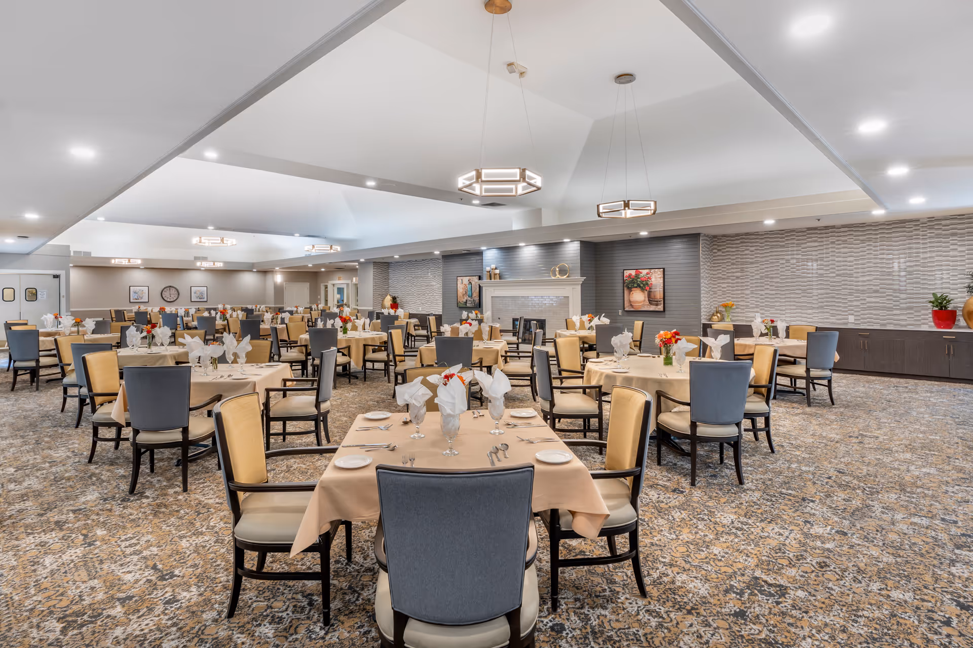 A spacious dining room with multiple round and rectangular tables covered with beige tablecloths, each set with plates, silverware, glasses, and napkins folded in an elegant design. The chairs have beige seats and backs with dark wooden frames. The room features a patterned carpet, modern pendant lights hanging from a high ceiling, and a decorative fireplace on the far wall with artwork and vases. The walls have textured paneling and there are flower arrangements on the tables and side cabinets.