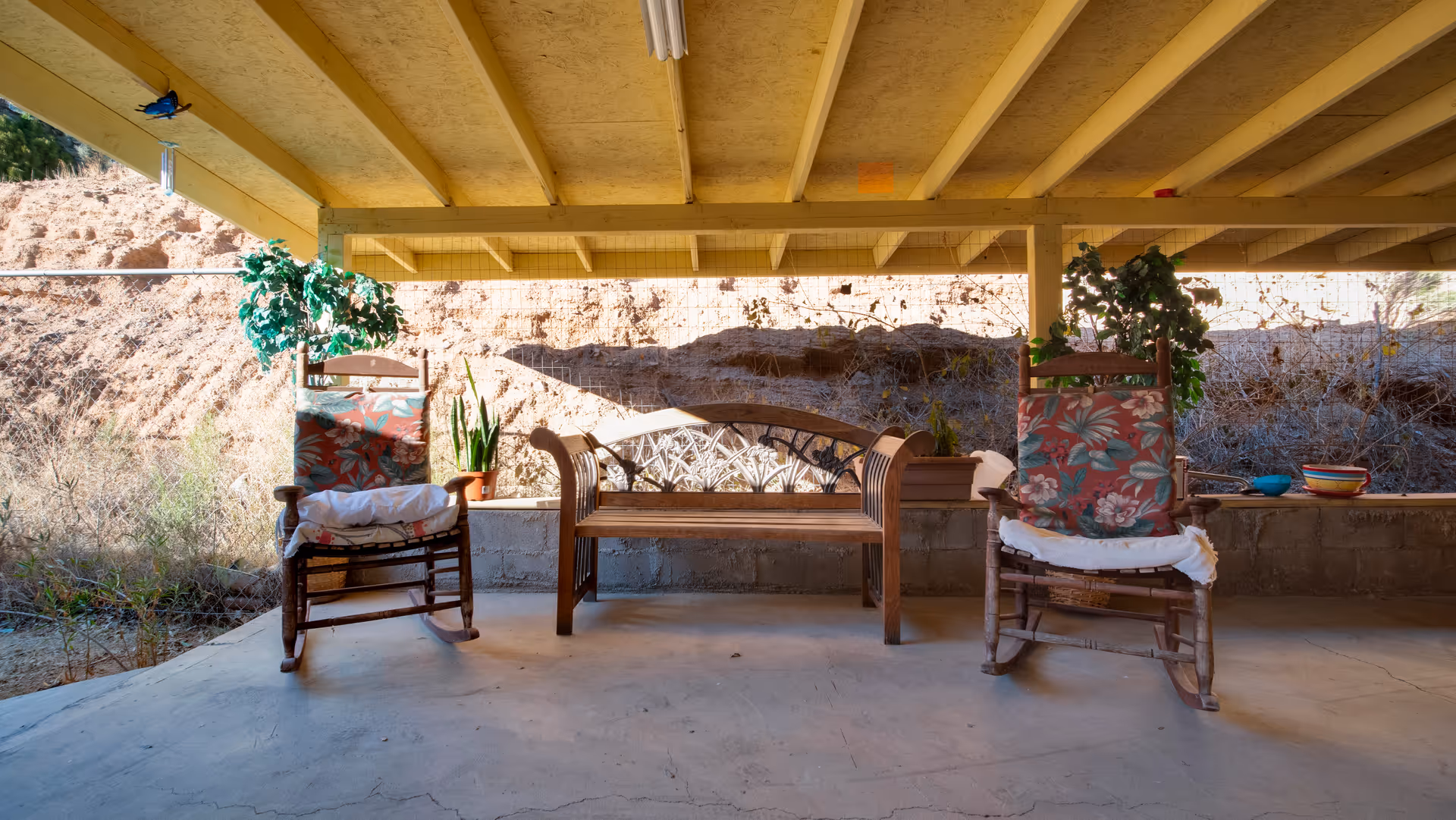 Covered outdoor patio area with two wooden rocking chairs with floral cushions and a wooden bench. There are potted plants on the ledge behind the seating, and a natural hillside is visible in the background.