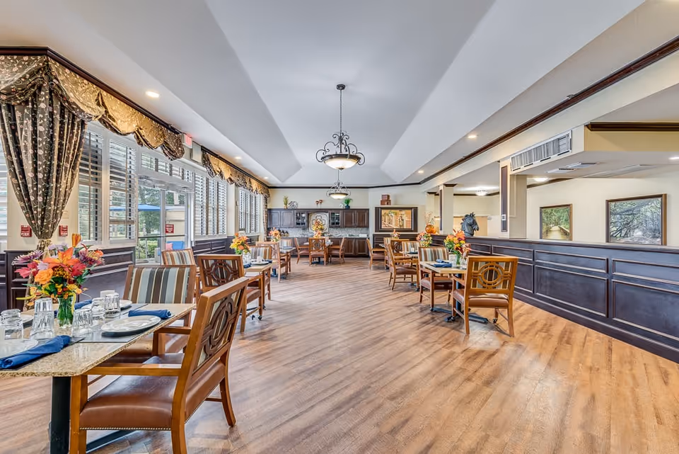 A bright and spacious dining room in a senior living facility with wooden floors, multiple tables set with plates, glasses, and napkins, floral centerpieces, large windows with patterned curtains, and decorative wall art.