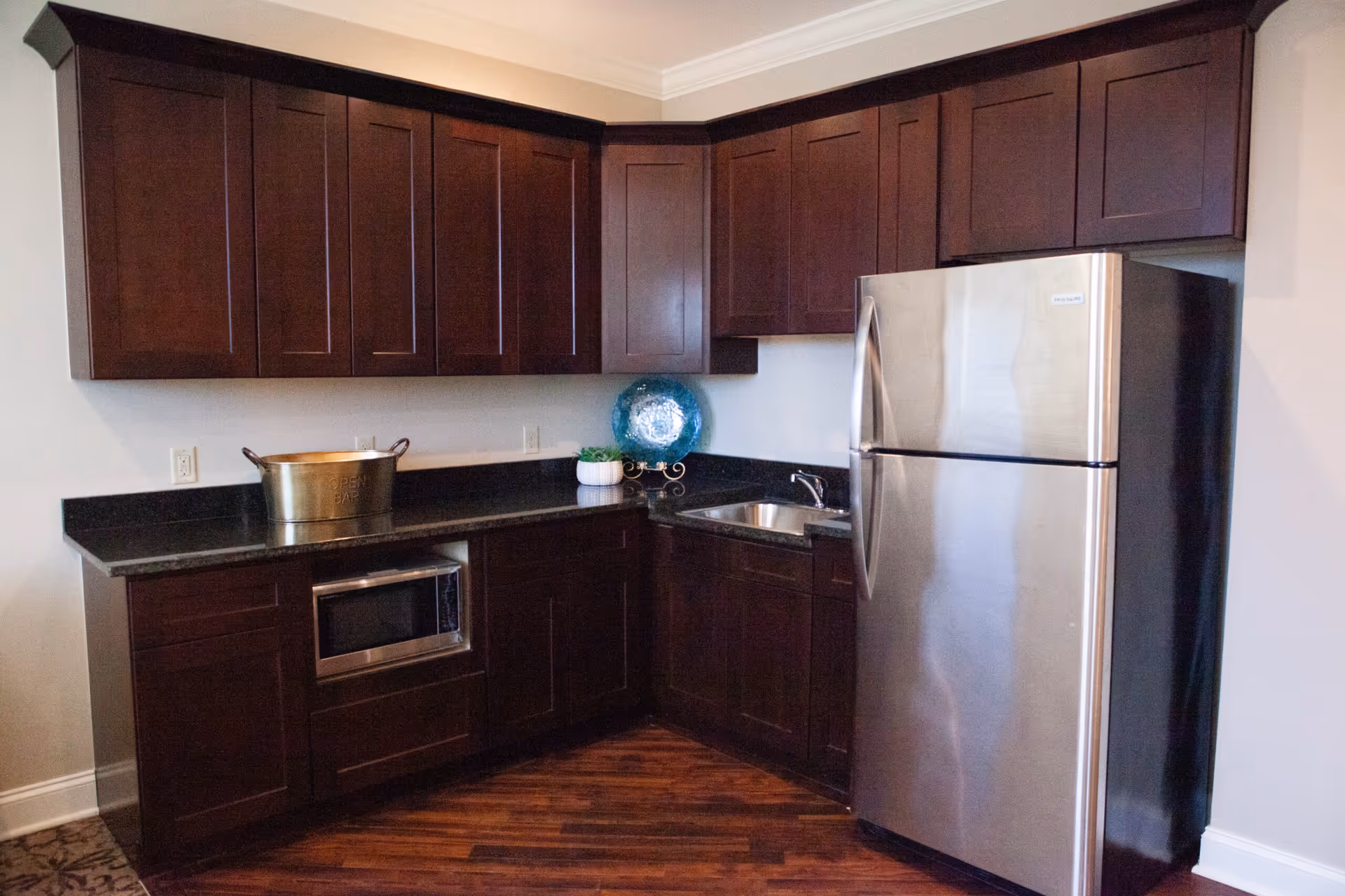 Small kitchen area with dark wood cabinets, granite countertops, a stainless steel refrigerator and a sink.
