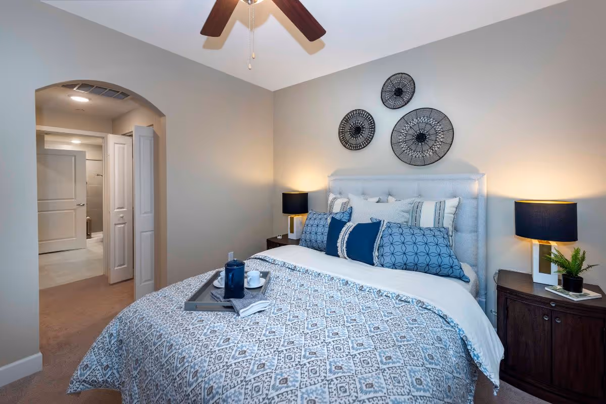 Neatly staged bedroom with a patterned bedspread, upholstered headboard, bedside tables with lamps, decorative wall art, and an arched doorway to an adjoining bathroom.
