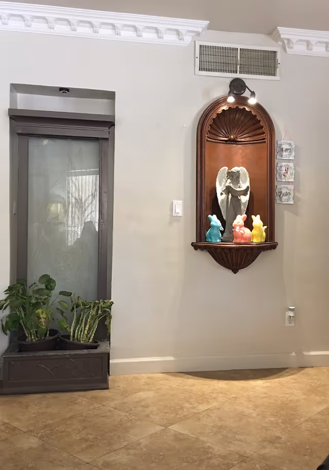 Interior wall with a decorative wooden niche holding a white angel statue and three colorful bunny figurines in blue, pink, and yellow. To the left, there is a small indoor planter with green plants. The wall is light-colored with crown molding at the ceiling and a vent above the niche. There is also a small hanging decoration with the words 'Happy Easter' on the right side of the niche.