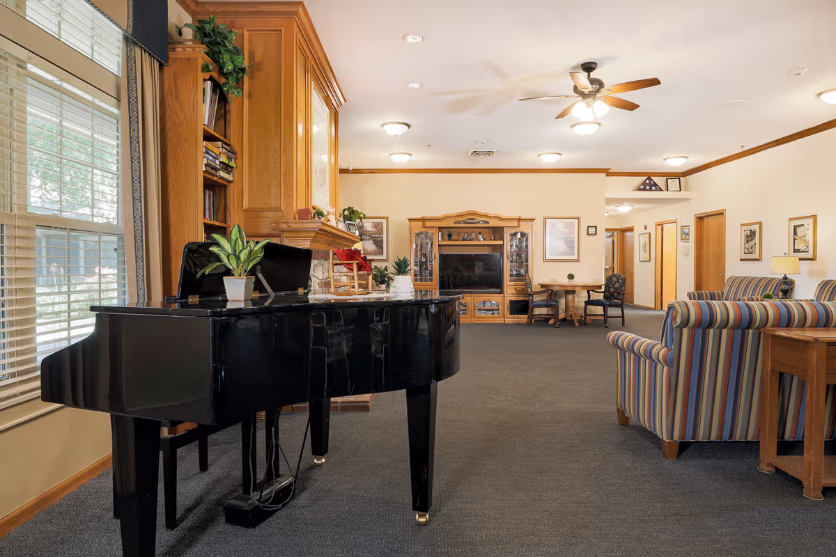 A spacious living room area in an assisted living facility featuring a black grand piano with a small potted plant on top, large windows with blinds, a wooden entertainment center with a TV, striped upholstered armchairs and sofas, a ceiling fan with lights, framed artwork on the walls, and a carpeted floor.