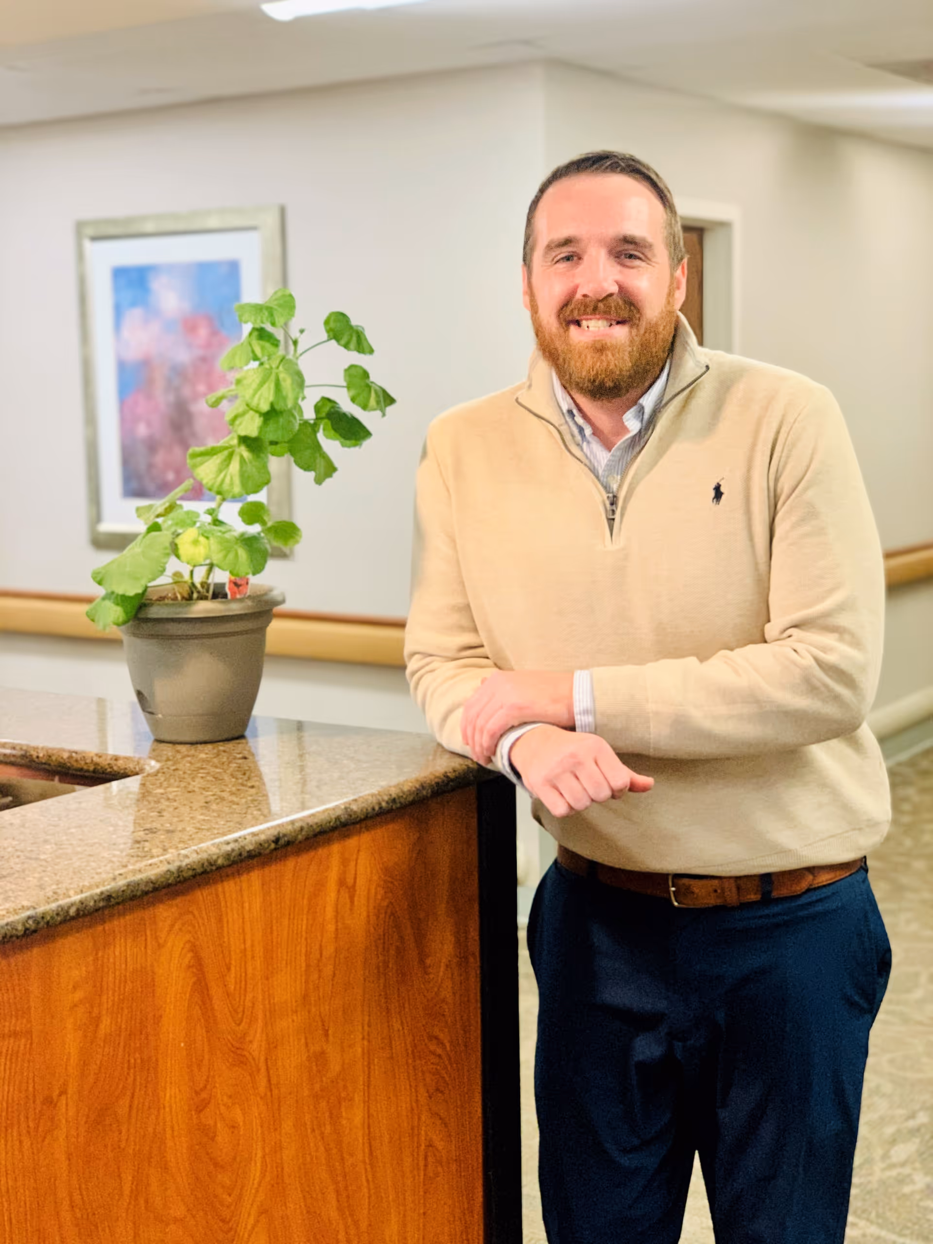 A man with a beard wearing a beige sweater and dark pants is standing and smiling while leaning on a wooden counter with a granite top. There is a potted plant on the counter and a framed picture on the wall behind him in a well-lit indoor setting.