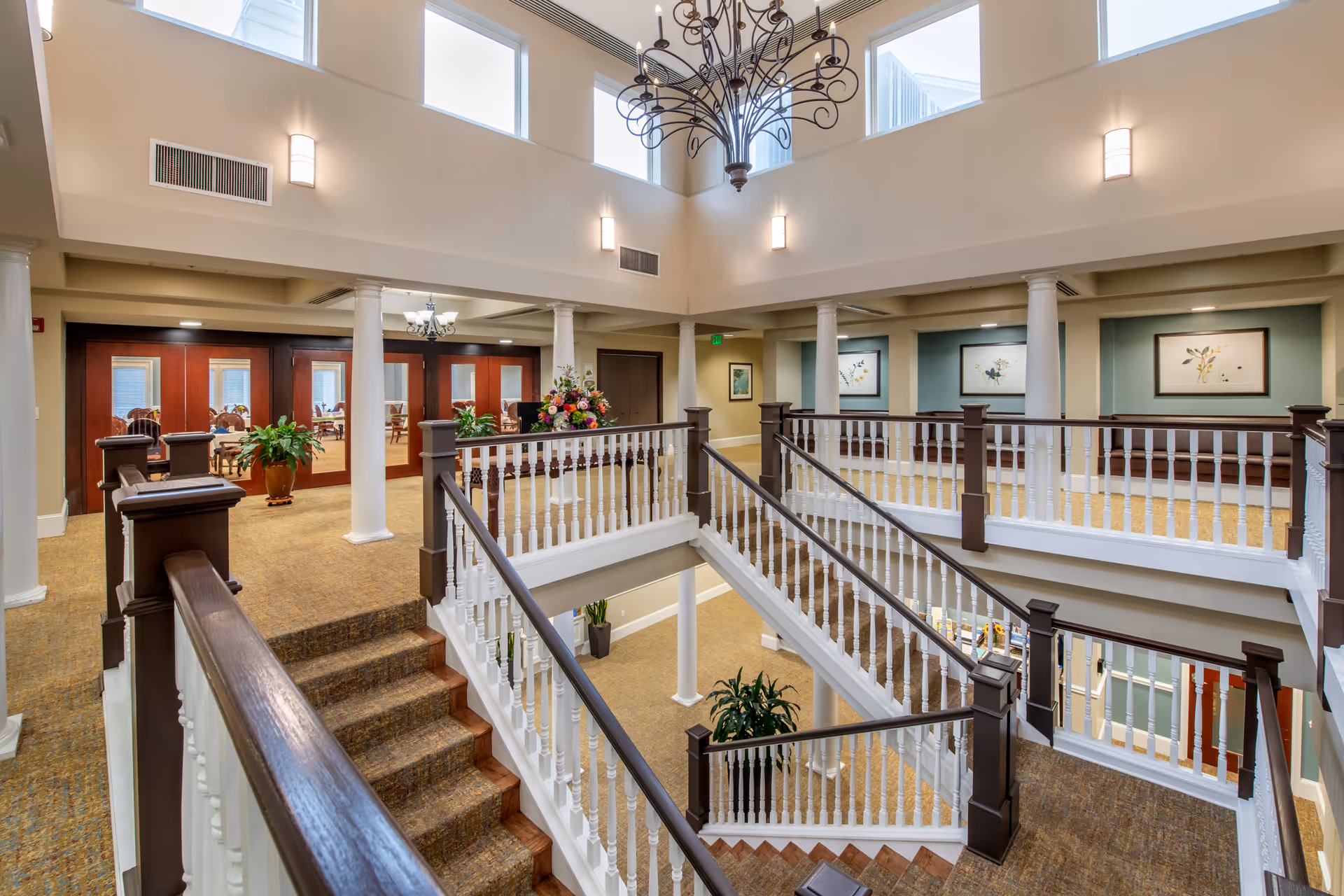 Interior view of a senior living facility showing a spacious two-story atrium with a staircase featuring white railings and dark wooden handrails. The upper level has framed artwork on the walls, potted plants, and a chandelier hanging from the ceiling. Large windows near the ceiling allow natural light to brighten the area.