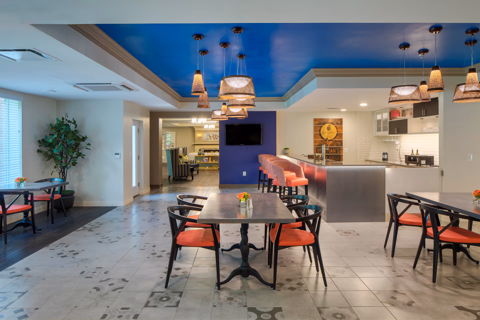 A modern dining area in a senior living facility with tables and chairs featuring red cushions. The room has a blue ceiling with multiple hanging pendant lights. There is a bar counter with pink bar stools and a wall-mounted TV. A plant is placed near the window on the left side, and the floor has a patterned tile design.