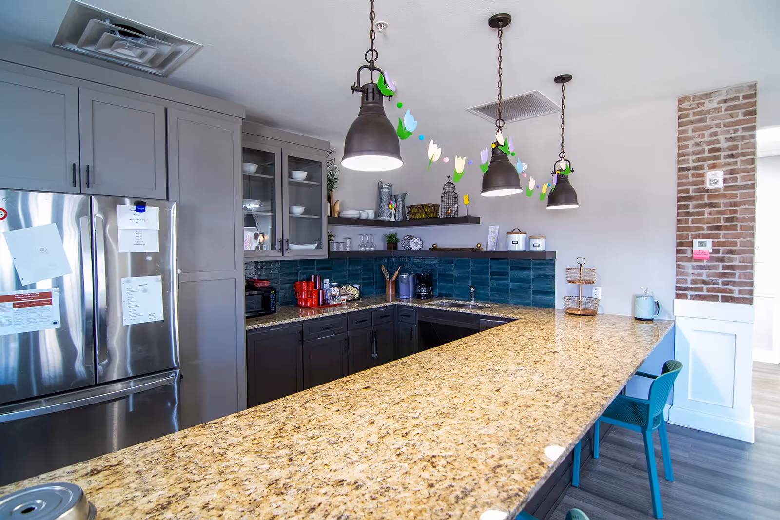 Modern kitchen area with granite countertops, dark lower cabinets, gray upper cabinets with glass doors, stainless steel refrigerator, three hanging pendant lights, and a decorative garland hanging from the ceiling. There are teal chairs tucked under the counter and a brick accent wall on the right side.