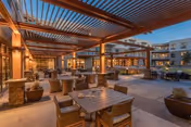 Outdoor patio area at dusk with wooden pergolas, multiple tables and chairs arranged for dining or socializing, large planters, and a multi-story building in the background with lit windows.