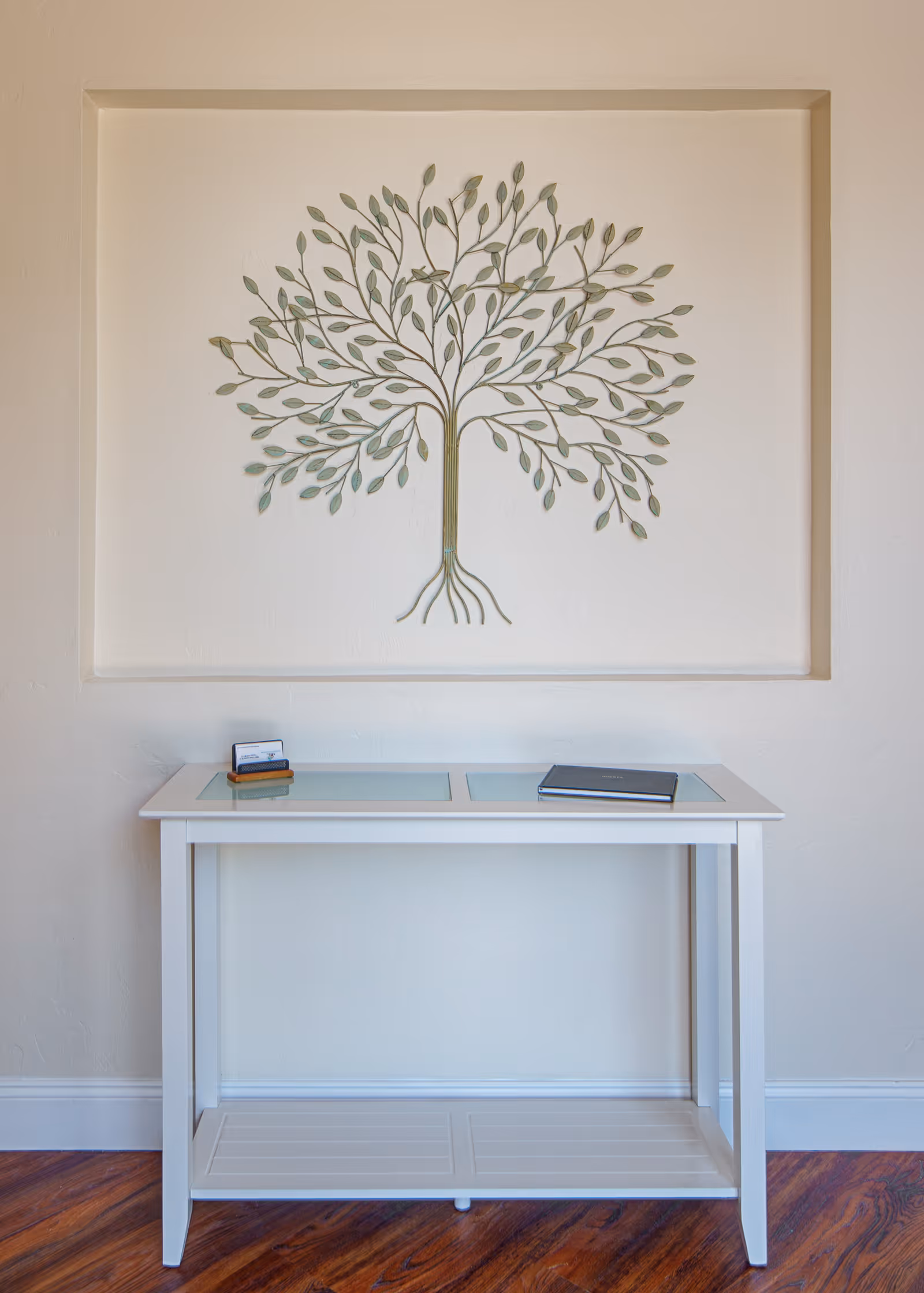 A white console table with glass panels on top, placed against a beige wall with a decorative metal tree sculpture mounted above it. The table holds a small stack of business cards and a closed book. The floor is wooden with a warm tone.
