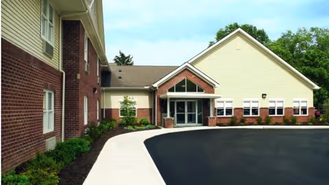 Exterior view of a single-story and two-story building with a brick and beige siding facade, featuring a main entrance with glass doors and a paved driveway leading up to it, surrounded by greenery and trees in the background.